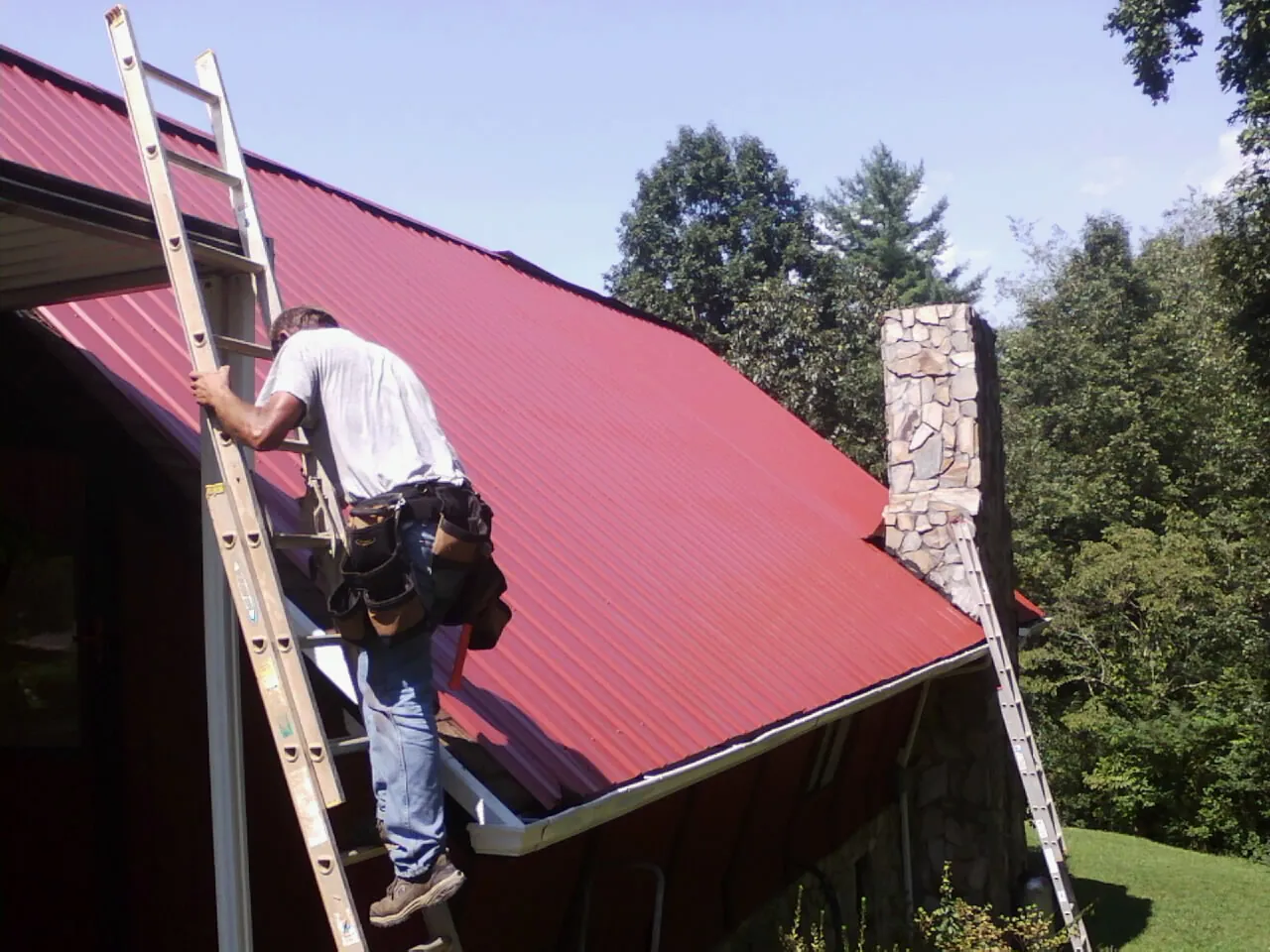 Roofer on a ladder beside a red metal roof and stone chimney — Soffit & Fascia Repair by Roofing Brothers in Cresco